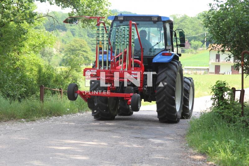 Traktor sõidab mööda maanteed, mille taga on punane põllumajandustootmise seade. Taimed ja rohelus ümbritsevad teed.