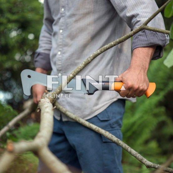 Mees hoiab käes saagi, millel on oranž käepide ja hõbedane tera. Ta seisab rohelise taimekeskkonna keskel ja lõikab ühte oksa.