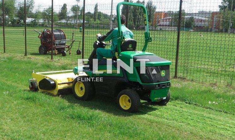 Muru niitja masin, millel on roheline ja must kere, niidab rohtu jalgpalliväljakul. Taustal on metallvõre ja niidukite käitamiseks mõeldud varustus.